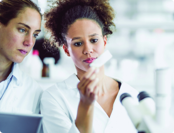 Two scientists in lab coats working together in a laboratory, reviewing information at a workstation with a microscope visible in the foreground